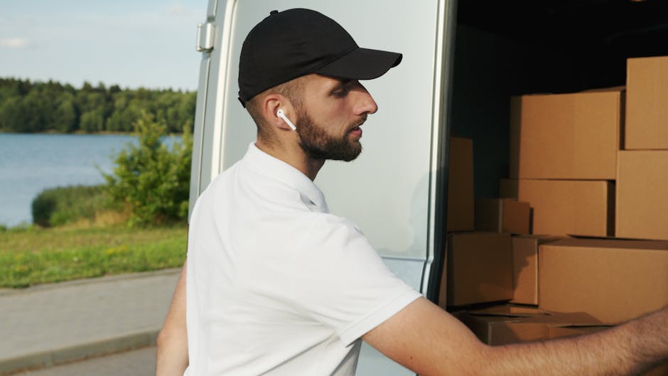 A male professional mover from Man and Van Bermondsey is standing outside a moving vehicle, ready to load or unload cardboard boxes that are stacked inside the van. He is wearing a white shirt, a blac