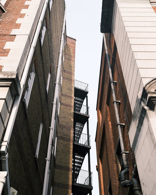 A view looking upward between two tall, closely situated buildings along a narrow street in Bermondsey, SE16. The building on the left has brickwork with white window frames and rain gutters running vertically down its side. The adjacent building on the right features an exterior clad in light-colored panels with decorative architectural elements and black metal downpipes. Between the buildings, a black metal fire escape with multiple landings and staircases is visible, extending upward along the façades. The sky above is partly visible, indicating daylight. This scene reflects the dense urban environment typical of Bermondsey, where house removals and furniture transport may involve navigating narrow streets and tight spaces, often requiring careful street-side loading processes, as demonstrated by the context of [COMPANY_NAME] providing removals services in such environments.