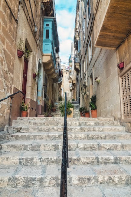 The image shows a narrow stone staircase leading upward through a residential area in Bermondsey, with various potted plants on either side placed on the steps and along the walls. To the left, there are multi-story houses with facades made of stone or brick, featuring small balconies and windows, some with flower boxes. On the right, a building with a stone exterior has an open window with a shutter and a small red wall-mounted light. At the top of the stairs, there are more buildings with mixed architectural styles, and a glimpse of the blue sky with scattered clouds is visible. A black metal handrail runs along the center of the staircase, aiding in the ascending movement, which is typical in close-knit residential areas like Bermondsey. This setting could serve as an environmentally authentic backdrop for themes related to house removals or home relocation, reflecting the tight urban streets and the logistical considerations of furniture transport and packing in such environments, as seen in services provided by Man and Van Bermondsey.