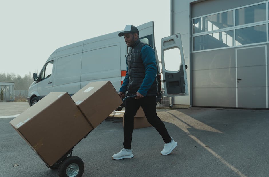 A man and a woman are engaged in a home relocation process, standing beside an open van loaded with numerous cardboard boxes of various sizes, some sealed with packing tape. The man, wearing a white p